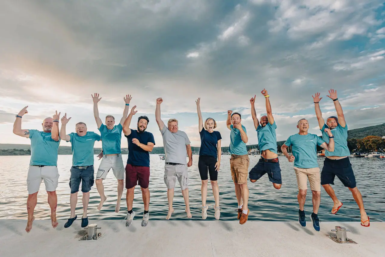 The In2theBlue crew in teal shirts jumping on the shore in front of the sea at Punat