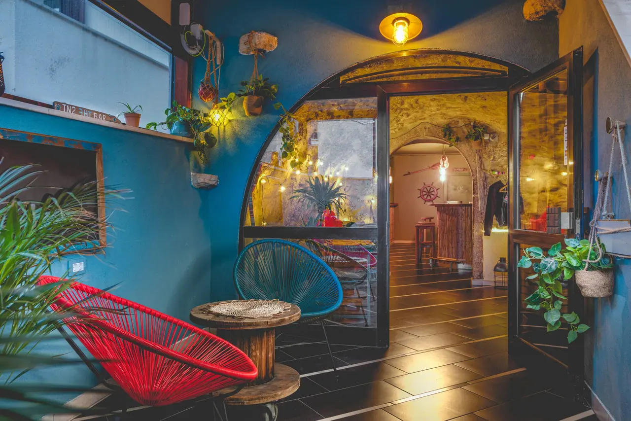 The arched entrance of In2 the bar, teal walls, a red Acapulco chair and warm lamps in the stone passage beyond