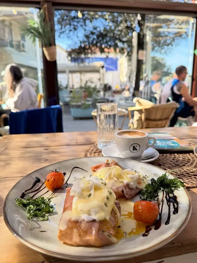 Eggs Benedict with prosciutto and hollandaise on a white plate, a coffee and the terrace behind it