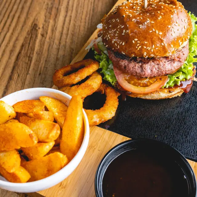 A beef burger in a sesame bun with lettuce and tomato on a slate board, beside a bowl of seasoned potato wedges, onion rings and a dip