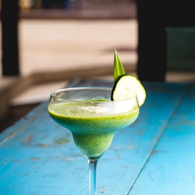 A pale green frozen drink in a coupe glass, garnished with a slice of cucumber