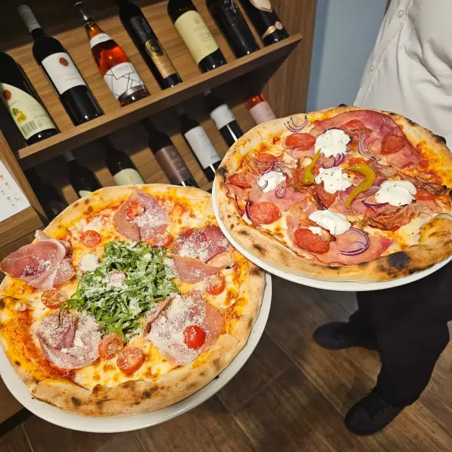 Two thin-crust pizzas carried out on plates, one with prosciutto, rocket and grated cheese, one with ham, salami, peppers and red onion