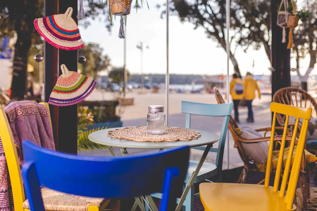 Blue and yellow chairs at a round table on the Punat waterfront promenade, straw hats hanging above