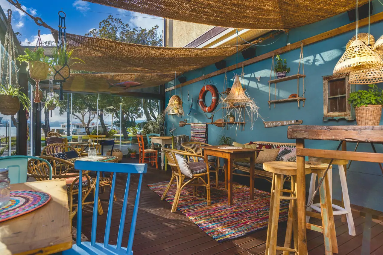 Teal terrace wall hung with oars, a life ring and shelves of plants, with rag rugs on the deck